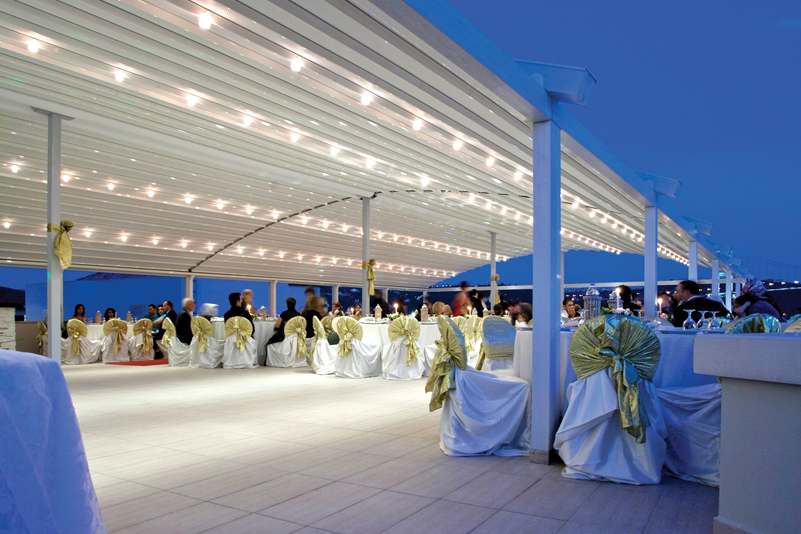 an open event area covered with a curved roof pergola with people inside sitting on chairs & chatting